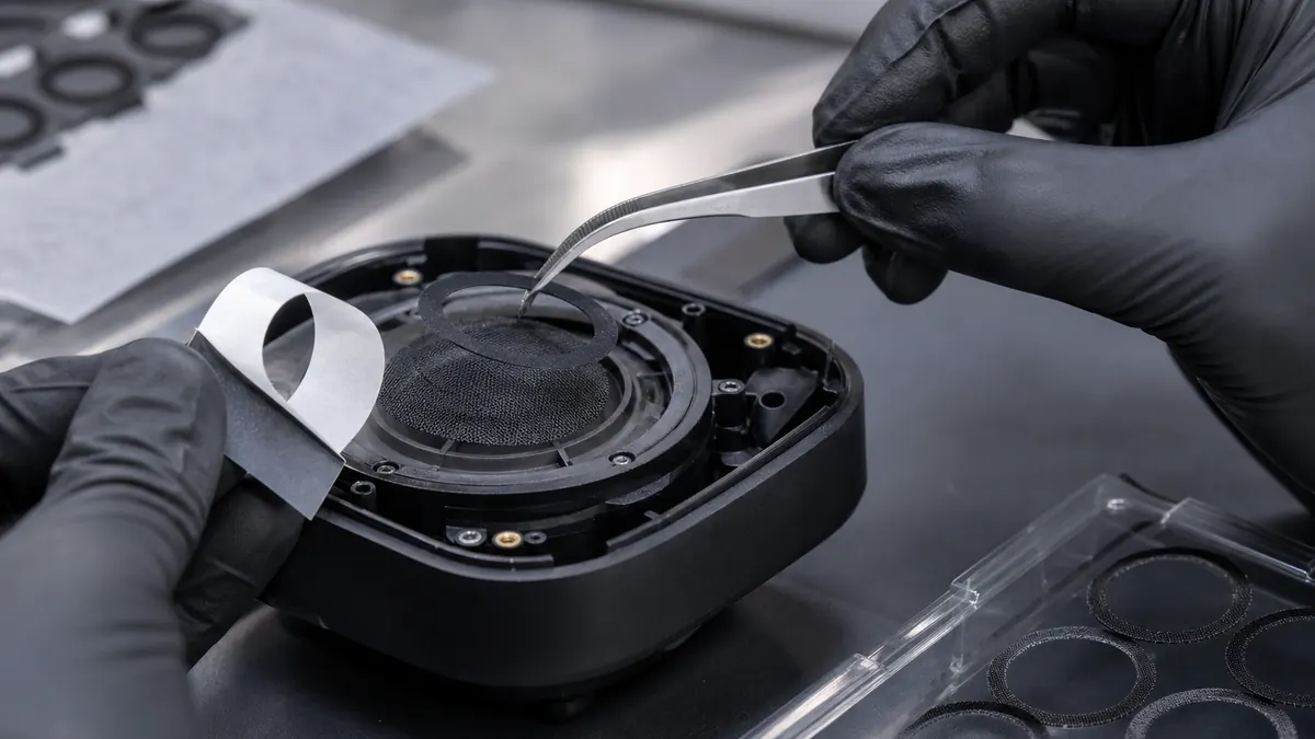 Technician aligning a black die-cut adhesive ring over speaker mesh in an electronics housing